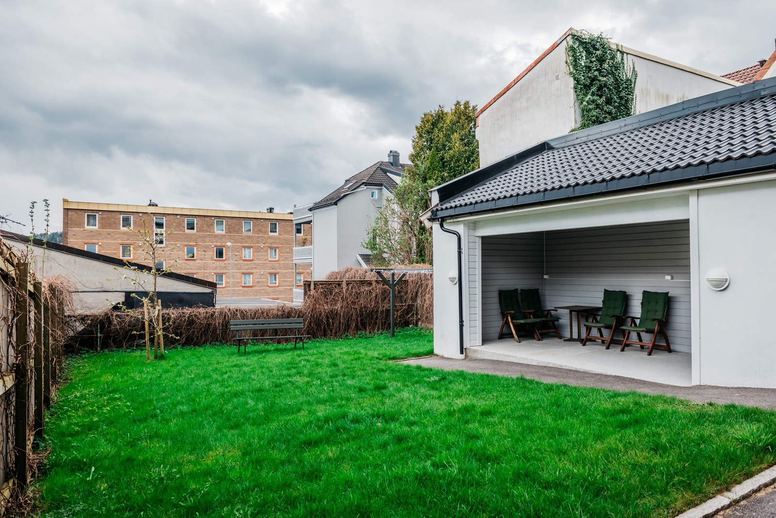 Fellesarealene er pent opparbeidet med asfaltert gangareal og stor gressplen, rammet inn av høye levegger.
Langs leveggene vokser klatreplanter som gir en grønn vegg om sommeren, og ved hagen er det etablert en åpen hagestue under tak med felles møbleme Galleribilde