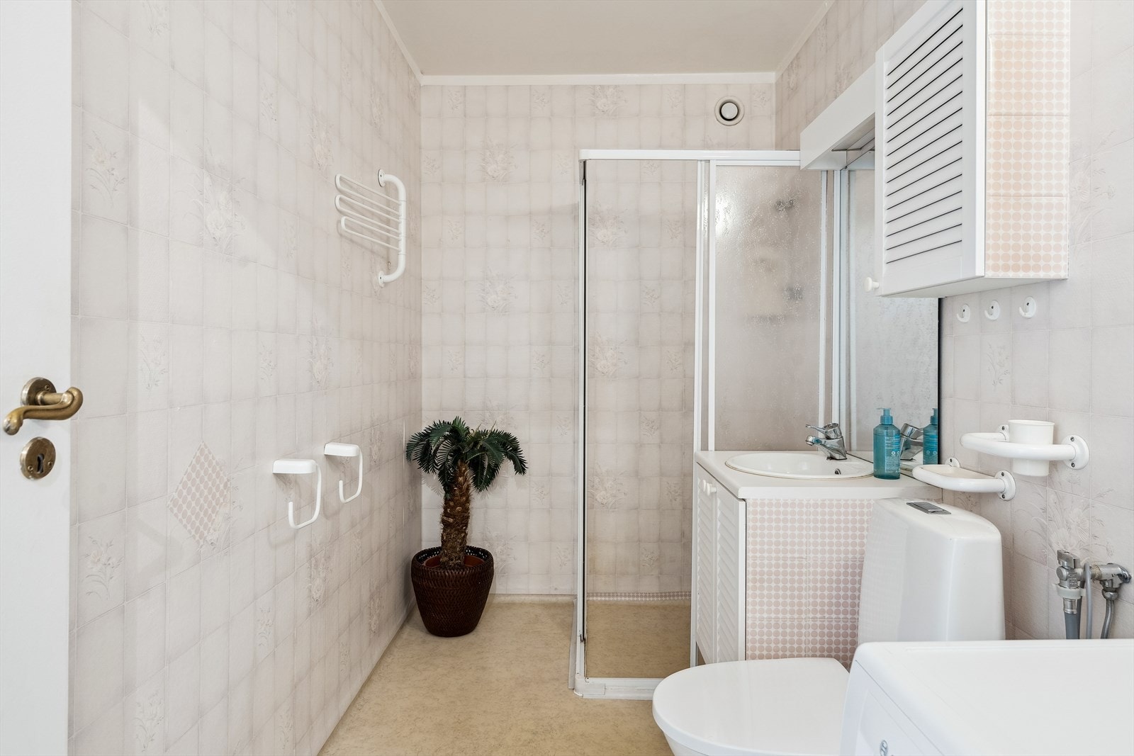 Eldre bad med gulvbelegg og vegger med vinyltapet. Badet er innredet med vask med skapinnredning, dusjhjørne med skyvedører og et toalett Galleribilde