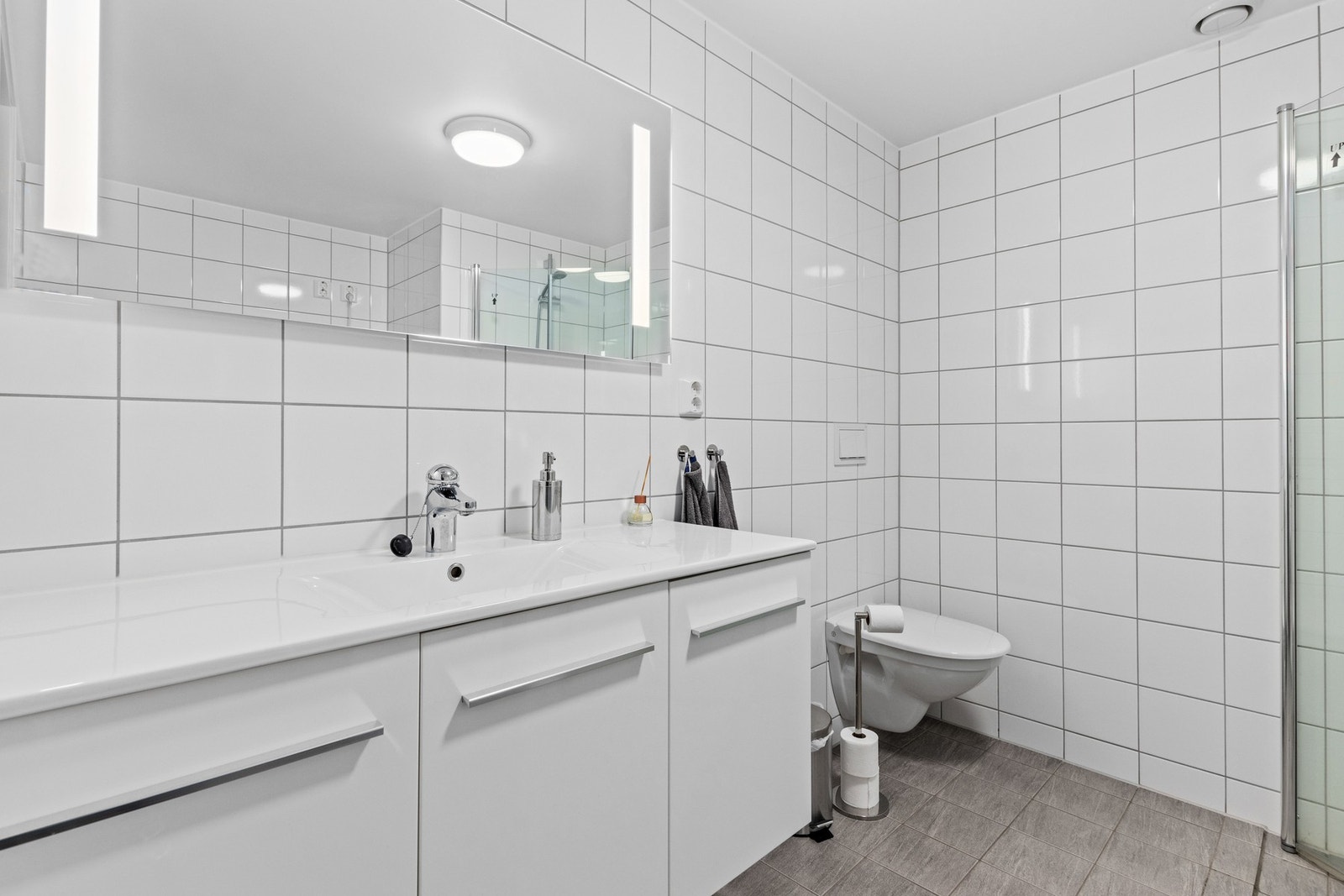Badet inneholder dusj med glass-dører, wc, servant i innredning, opplegg for vaskemaskin og en utslagsvask. Galleribilde