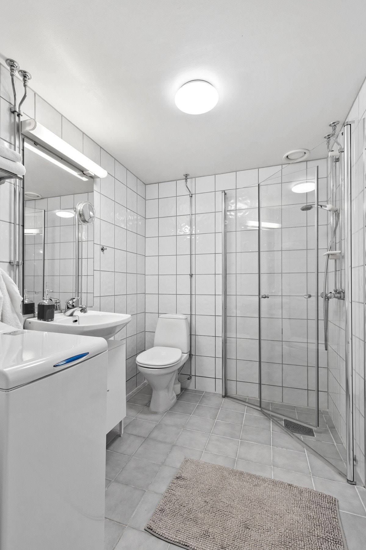 Badet har hvit servantinnredning, dusjhjørne med glassdører og vegghengt dusjarmatur, samt gulvstående toalett Galleribilde