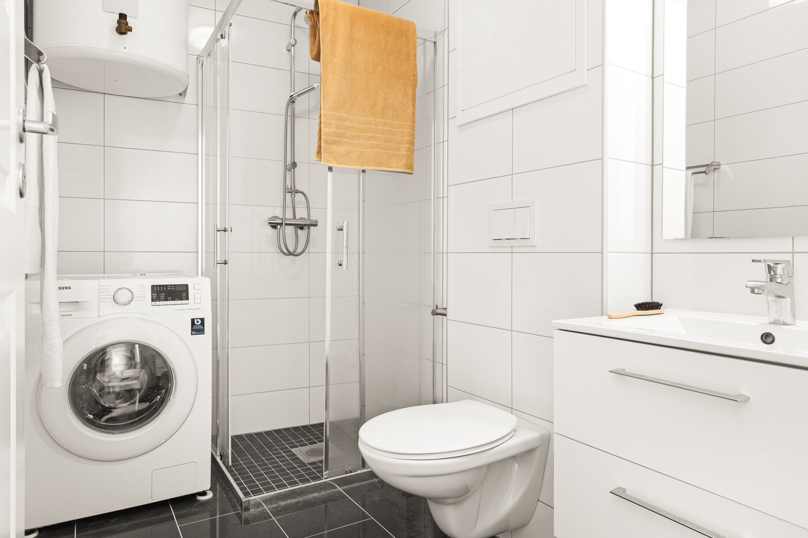 Badet er helfliset og inneholder dusjhjørne, vegghengt toalett, baderomsinnredning og opplegg for vaskemaskin. Galleribilde