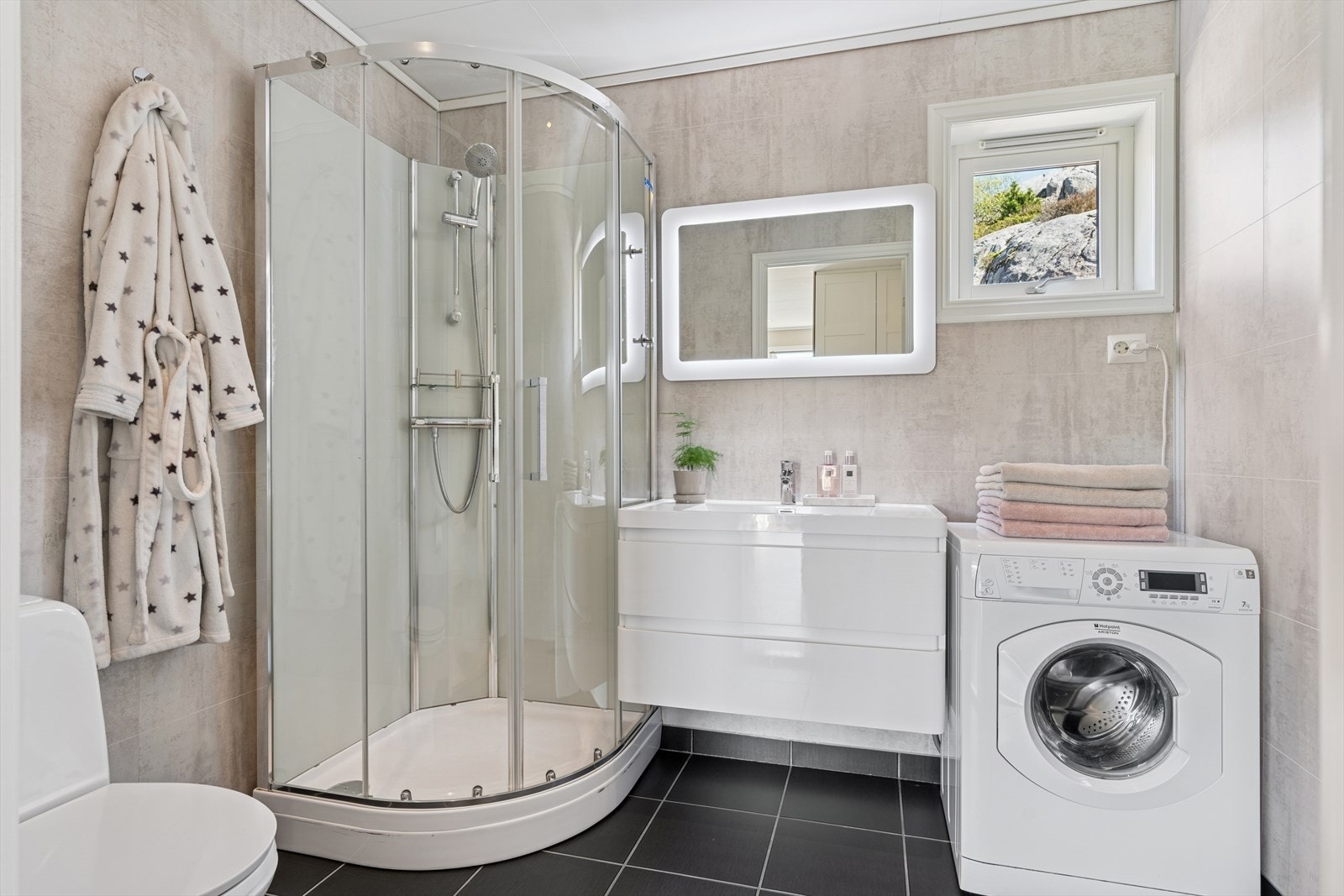 Innbydende bad/vaskerom med fliser på gulv og våtromsplater på vegger. Badet fremstår som en romslig velværeavdeling, for her bys det på dusjkabinett, toalett, seksjon med servant og opplegg for vaskemaskin. Avtrekk via mekanisk avtrekksanlegg. Galleribilde