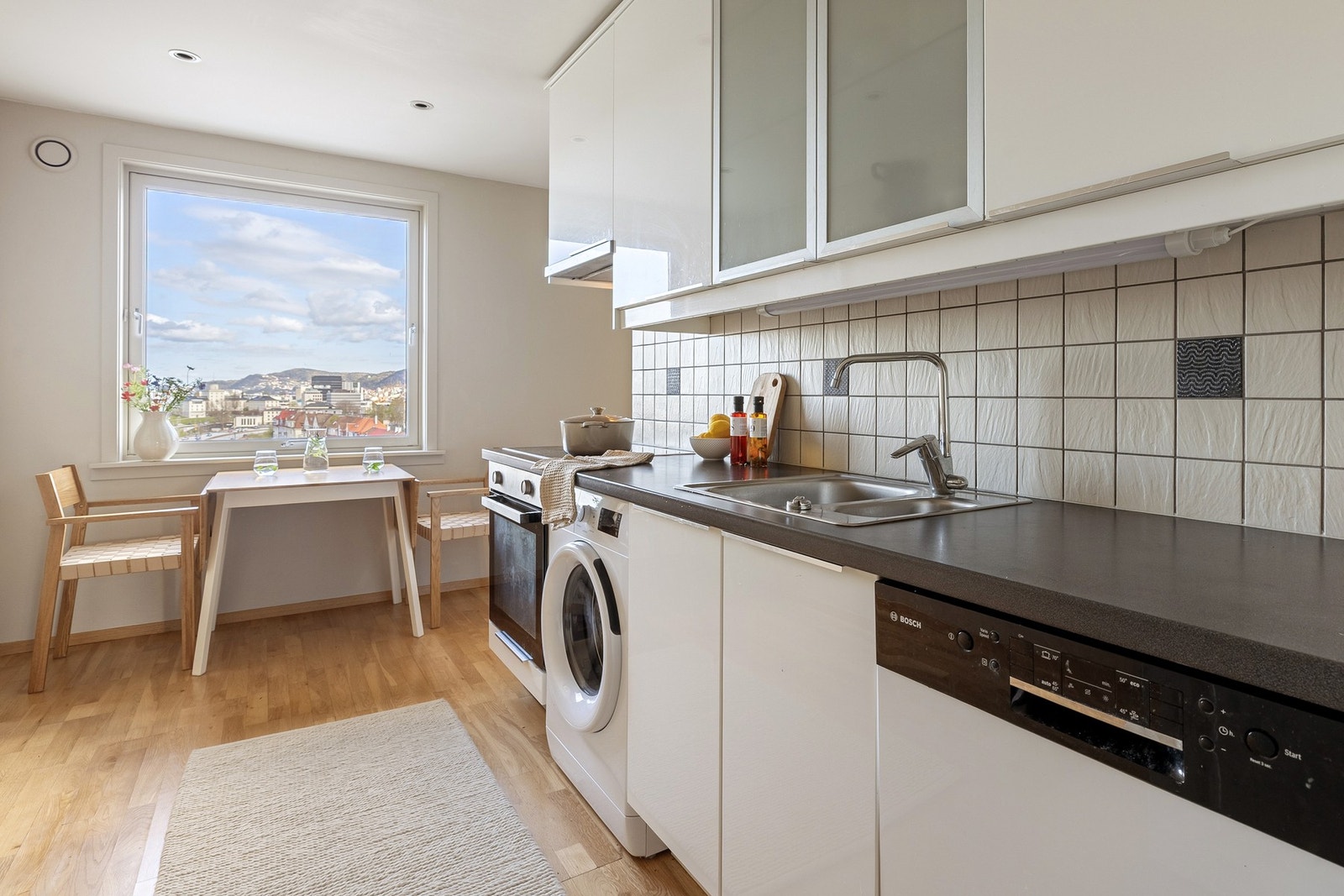 Kjøkkeninnredning består av hvite, slette fronter, to glasskap og laminat benkeplate med nedfelt vask av stål. Galleribilde