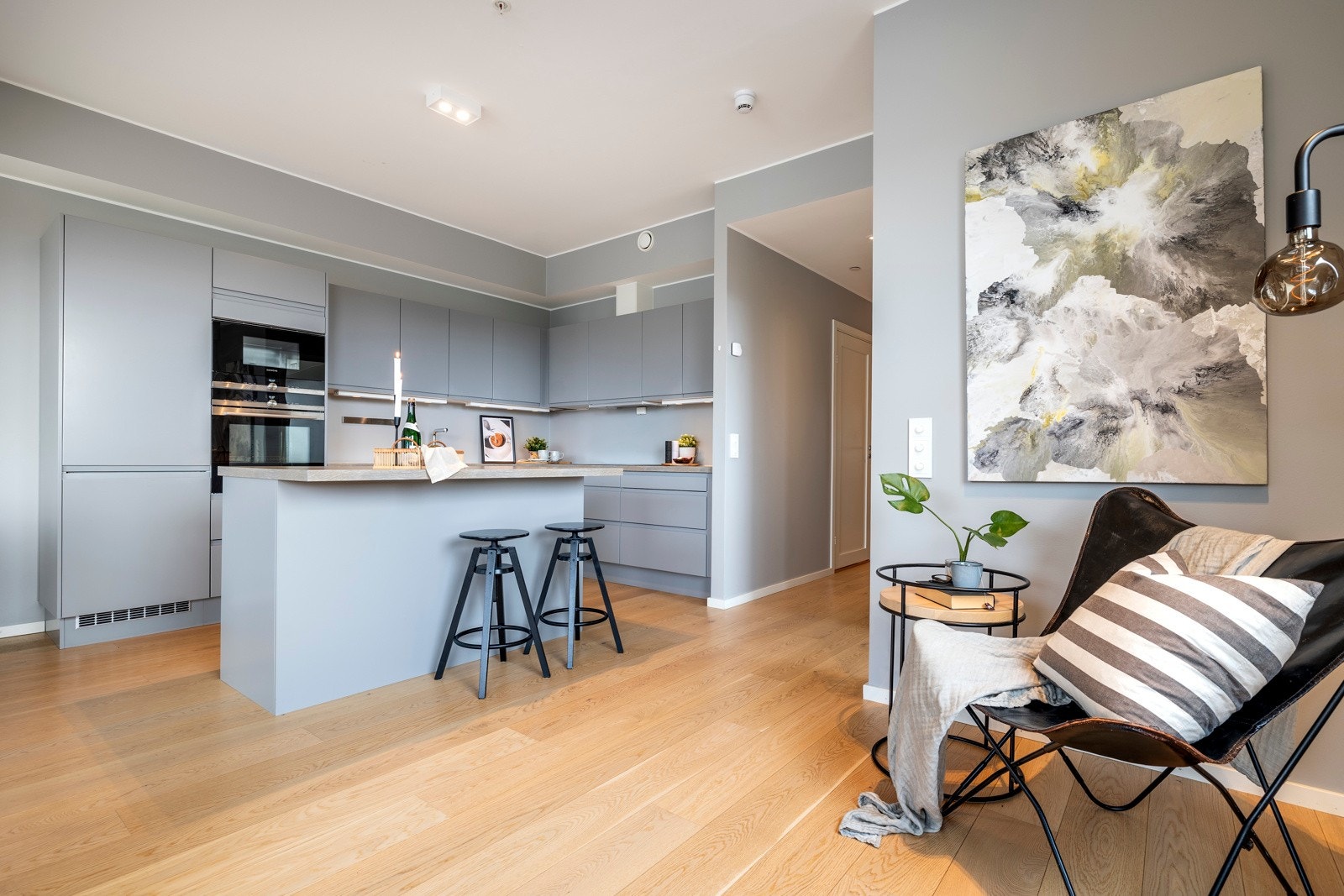 Kjøkkenet er plassert i en hendig L-form, innerst i stuen, pluss en stor øy med barløsningen i overgangen til stuesonen. Innredningen har glatte, grå fronter, nært opp til veggfargen, slik at den visuelt nærmest går i ett med bakgrunnen. Galleribilde