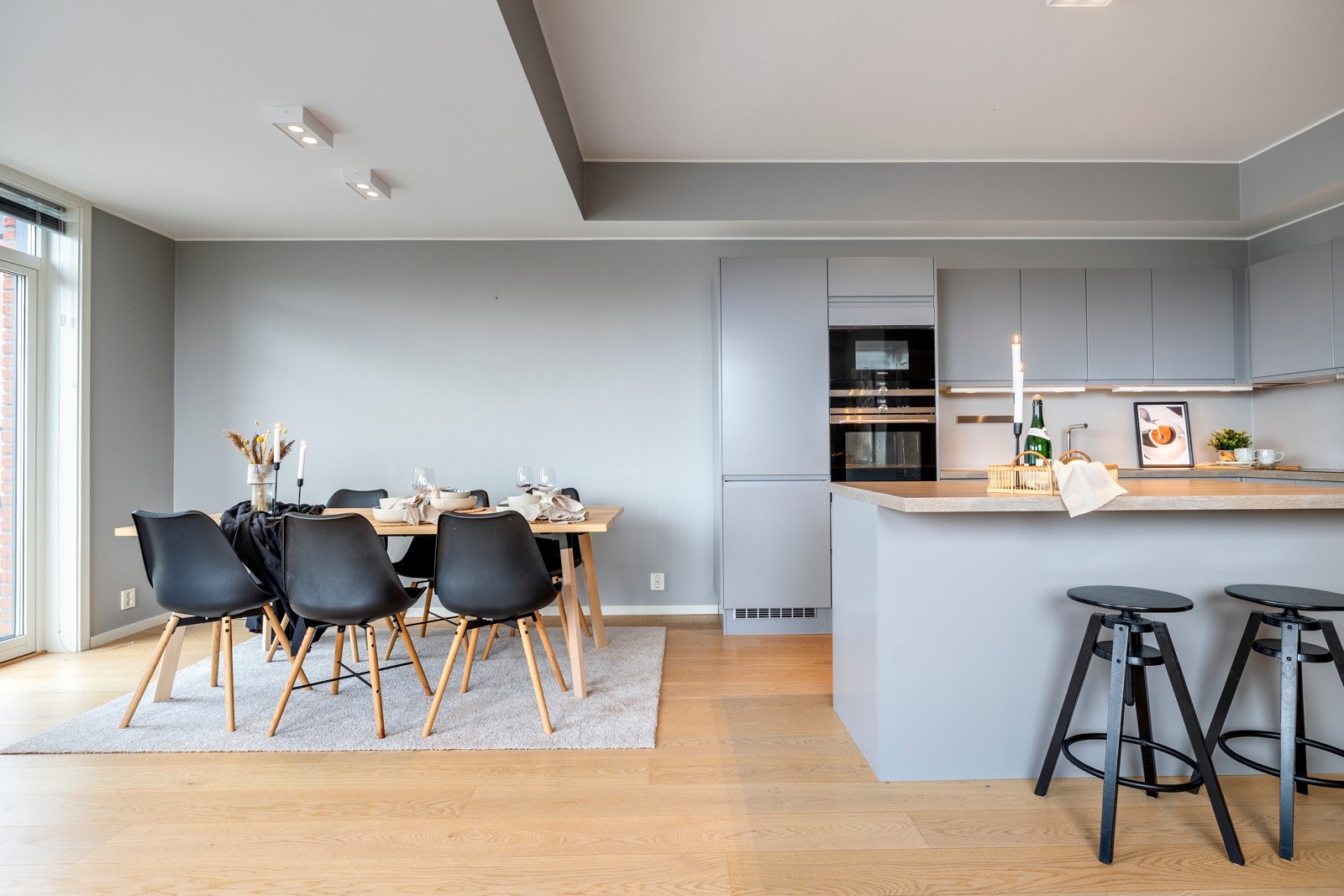 Kjøkkenet er plassert i en hendig L-form, innerst i stuen, pluss en stor øy med barløsningen i overgangen til stuesonen. Innredningen har glatte, grå fronter, nært opp til veggfargen, slik at den visuelt nærmest går i ett med bakgrunnen. Galleribilde