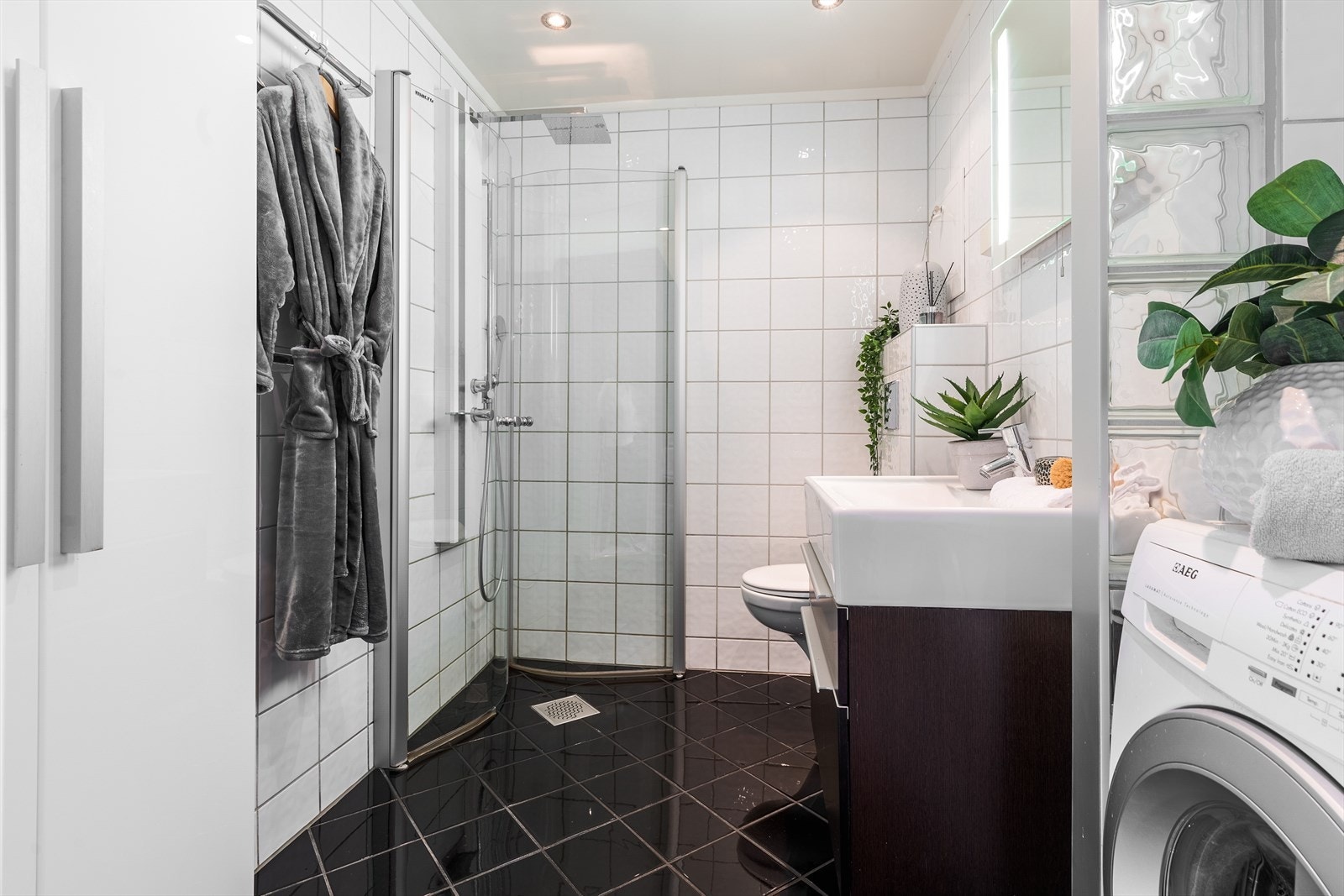 Badet er innredet med servant på skuffeseksjon, vegghengt toalett, dushjørne og opplegg for vaskemaskin. Galleribilde