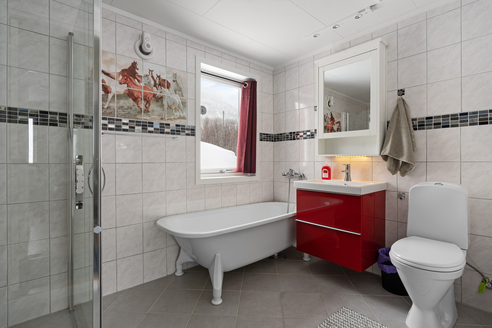 Badet er utstrt med badekar, dusjhjørne, toalett og vegghengt servant med oppbevaring i skuffer.
