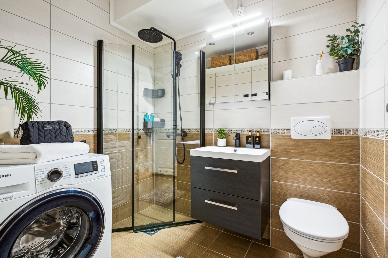 Stilrent baderom utstyrt med moderne servantinnredning, speilskap, dusjhjørne med innfellbare dører av glass, vegghengt toalett og opplegg for vaskemaskin. Galleribilde