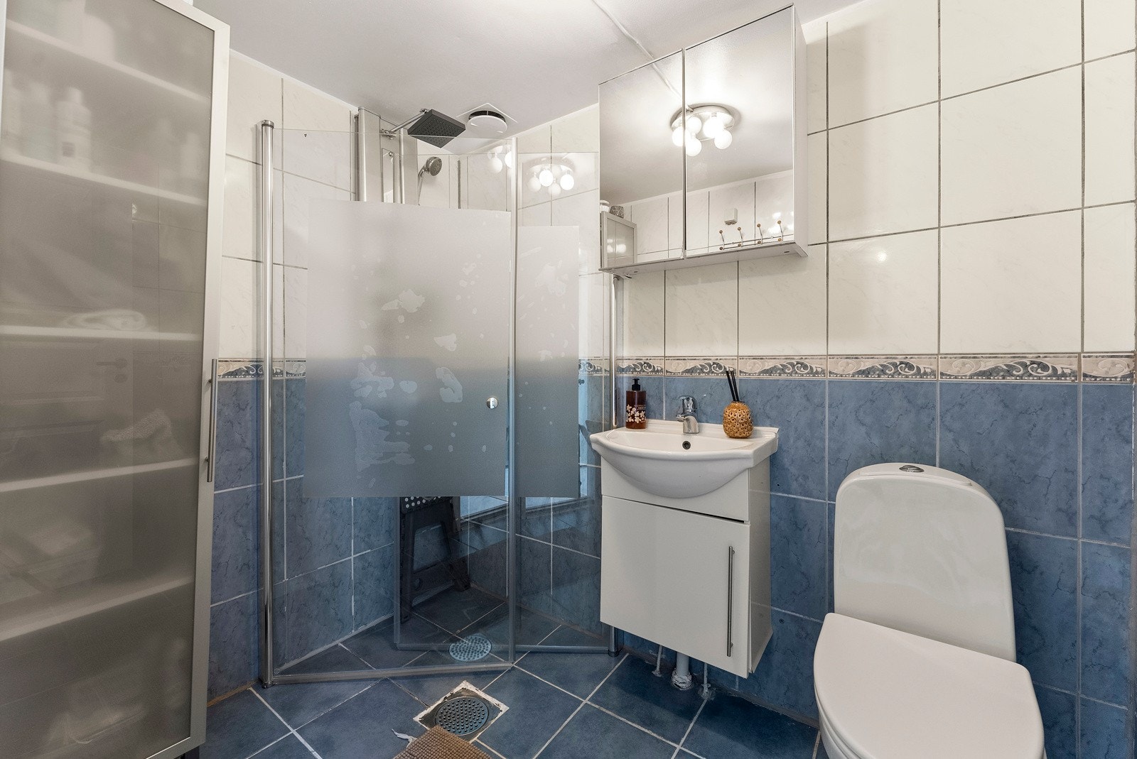 Badet er romslig og har fliser på gulv og vegger. Det er utstyrt med gulvmontert toalett, nedsenket servant med innredning og speil, samt en dusjnisje med glassdører. Galleribilde