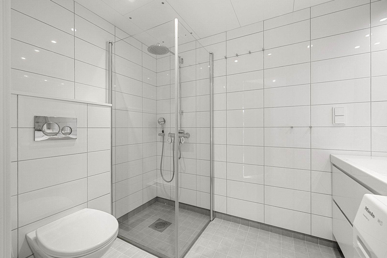 Badet er utstyrt med vegghengt toalett, servant med skuffer og dusjnisje med innfellbare glassdører. Galleribilde