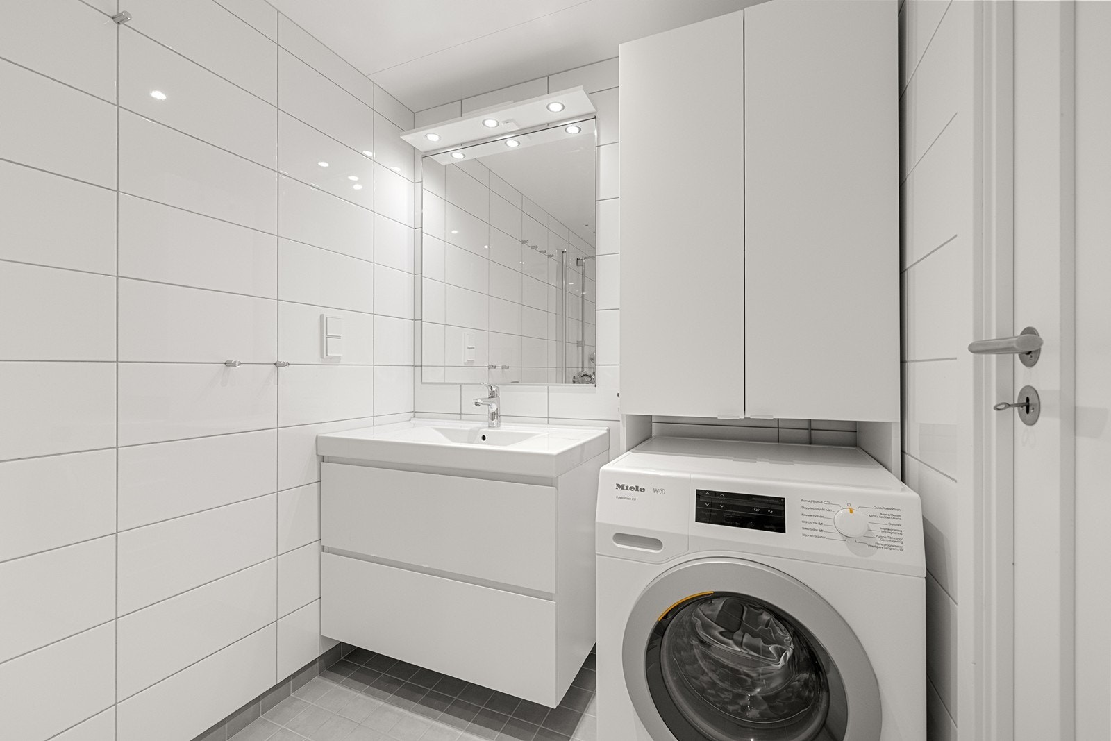 Pent flislagt baderom i tidløse fargetoner. Speil med belysning i overkant av servant og oppbevaringsskap på vegg. Opplegg for vaskemaskin og vannbåren varme i gulv. Galleribilde