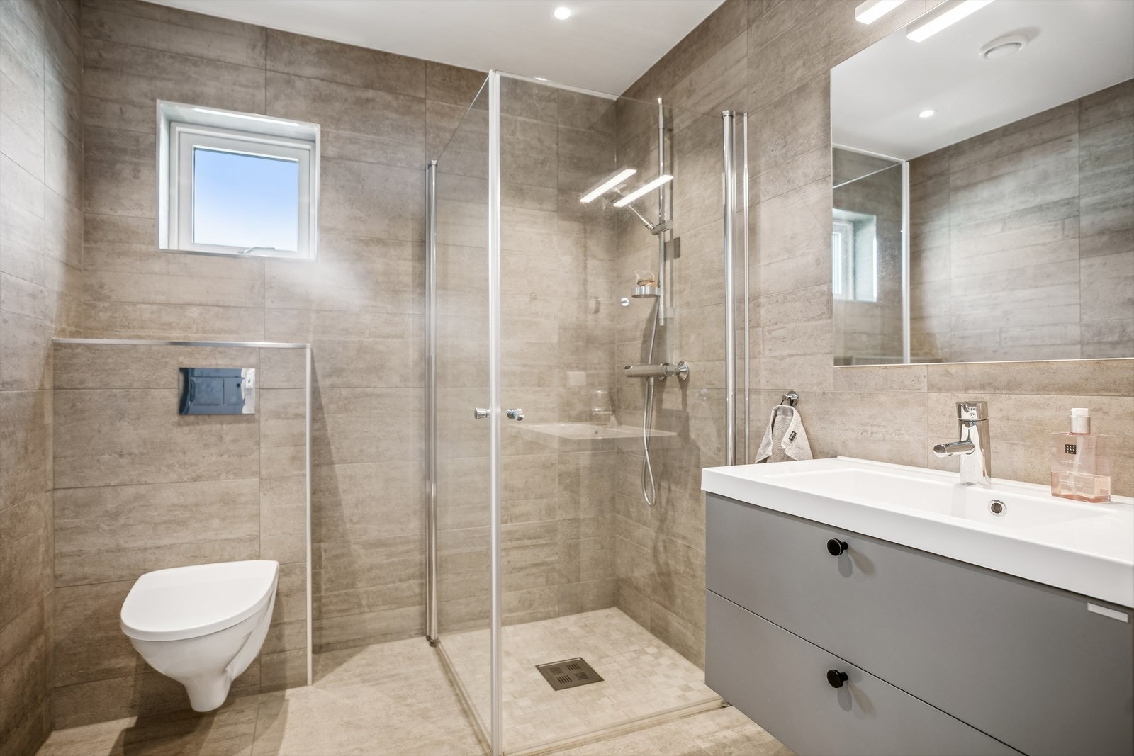 Lekkert og flislagt baderom med dusjhjørne, vegghengt toalett, servant med underskap og speil. Galleribilde