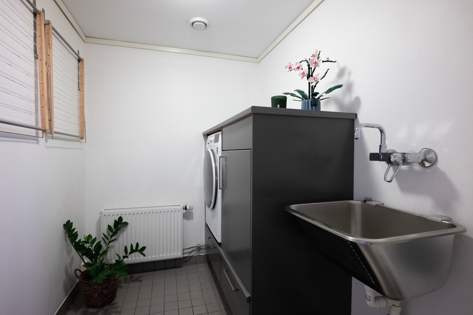 Flislagt vaskerom med vegghengt utslagsvask og gulvstående benkeskap med opplegg for vaskemaskin. Vaskerommet bidrar til en enklere hverdagslogistikk for hele familien. Galleribilde