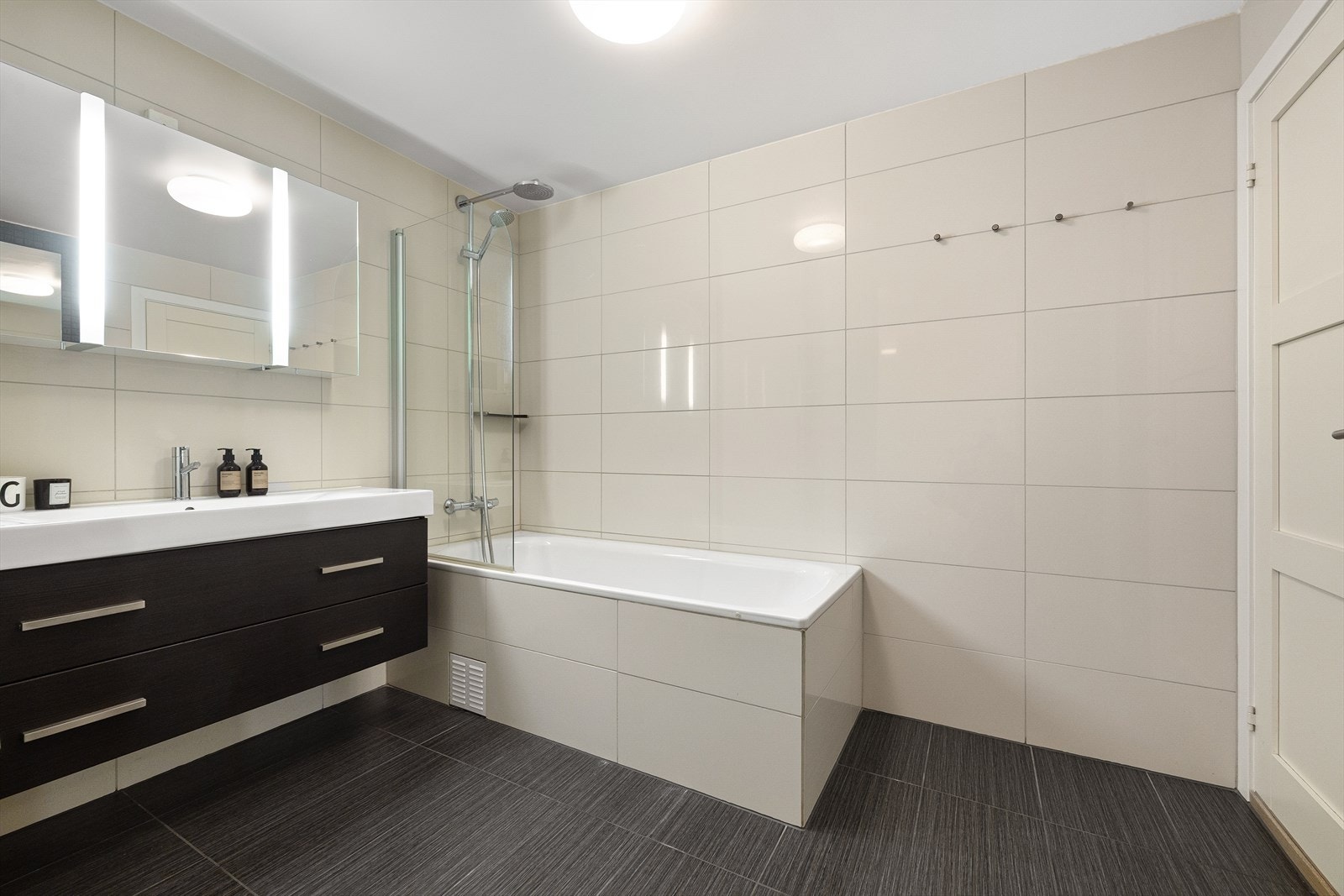 Badet er innredet med servant i innredning med skuffer, speilskap med belysning, badekar med glassvegg og dusjløsning, skap på vegg, samt vegghengt toalett Galleribilde