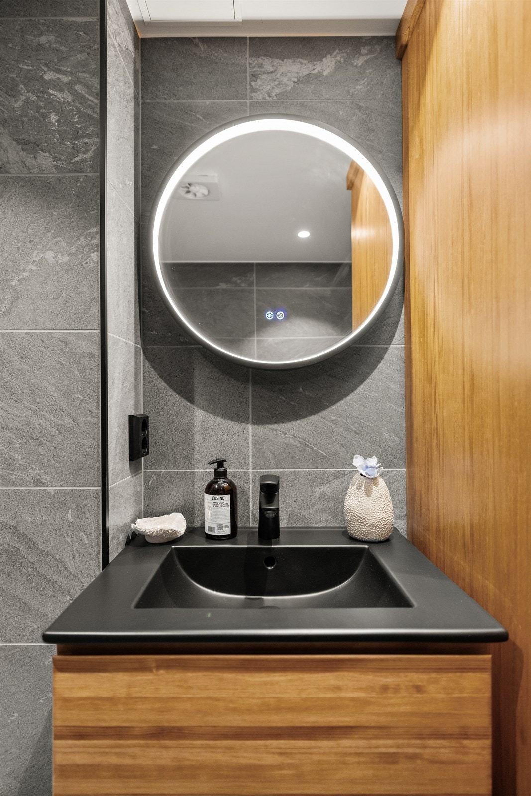 Badet er innredet med sort servant, trehvite fronter, rundt speil og dusjhjørne med regnfallsdusj. Galleribilde