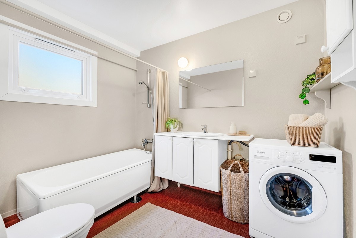 Badet er utstyrt med servantinnredning, speil, badekar med dusjgarnityr, gulvmontert toalett og opplegg for vaskemaskin Galleribilde