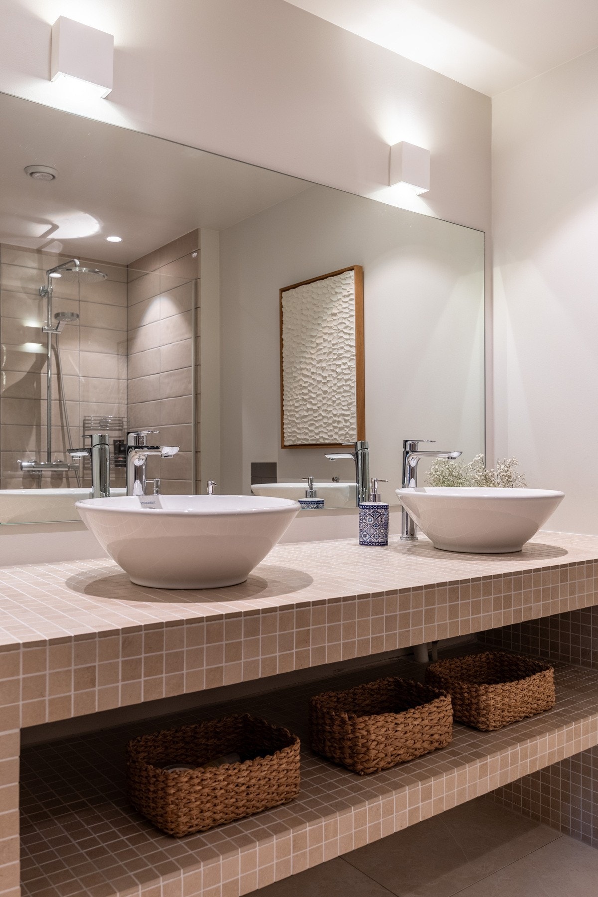 Dette badet har lyse gulvfliser, hvite vegger, lekre glaserte veggfliser i dusjsonen, downlights og gulvvarme. Innredningen består av to bolleservanter på en mosaikkledt servantbenk, stort speil med belysning og et veggmontert toalett. Galleribilde