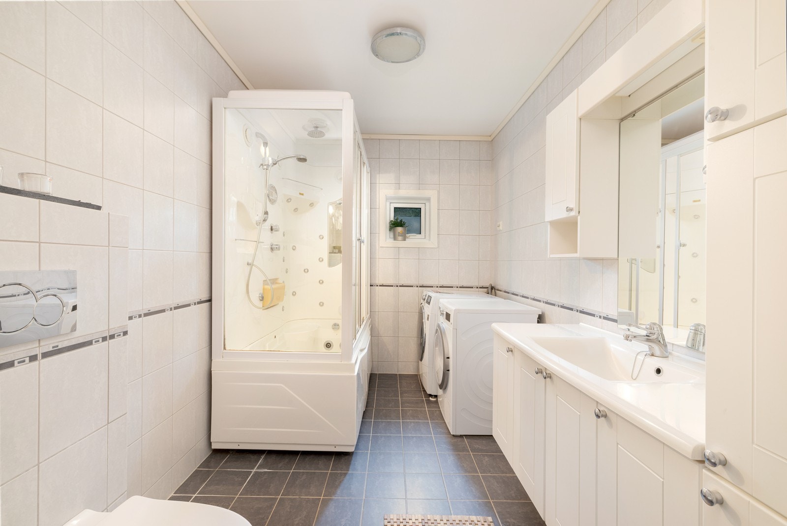 Flislagt bad med varme i gulv. Badet har et stort dusjkabinett med flere funksjoner, vegghengt toalett, servant med underskap og opplegg til vaskemaskin og tørketrommel. Galleribilde