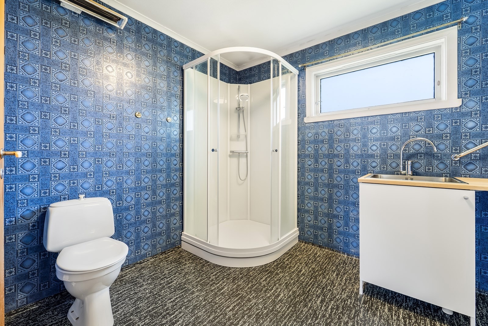 Eldre bad med belegg på gulv og tapet på vegg. Innredet med baderomsinnredning, toalett, dusjkabinett og en vaskeromsavdeling med opplegg til vaskemaskin. Galleribilde