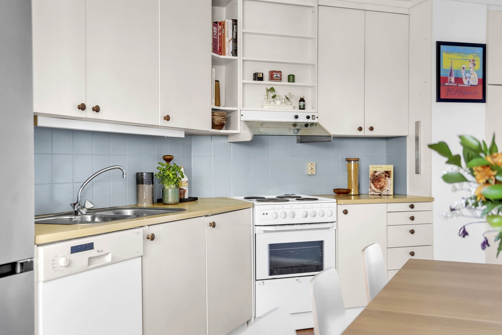 Frittstående kjøleskap med frysedel og frittstående komfyr med kokeplater. Det er ventilator under overskap og oppvaskmaskin under benk. Galleribilde