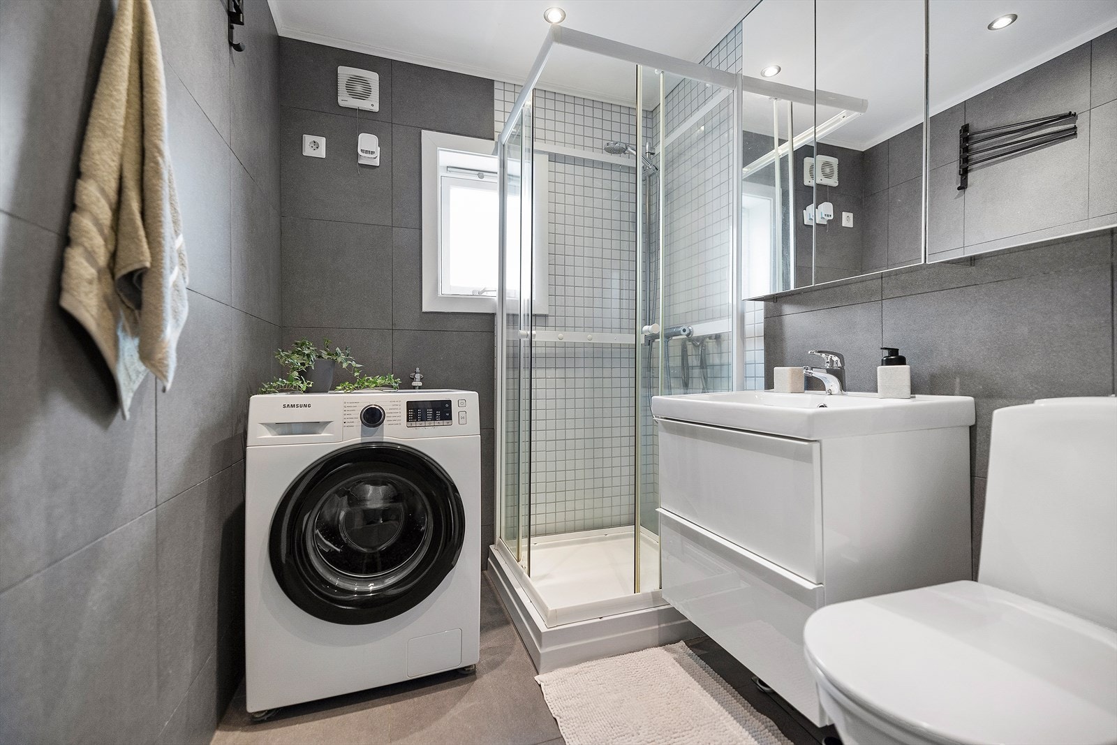 Badet er flislagt og har elektriske varmekabler i gulvet. Innredningen består av en nedfelt servant, vegghengt speilskap, gulvmontert toalett, dusjkabinett og opplegg for vaskemaskin. Galleribilde