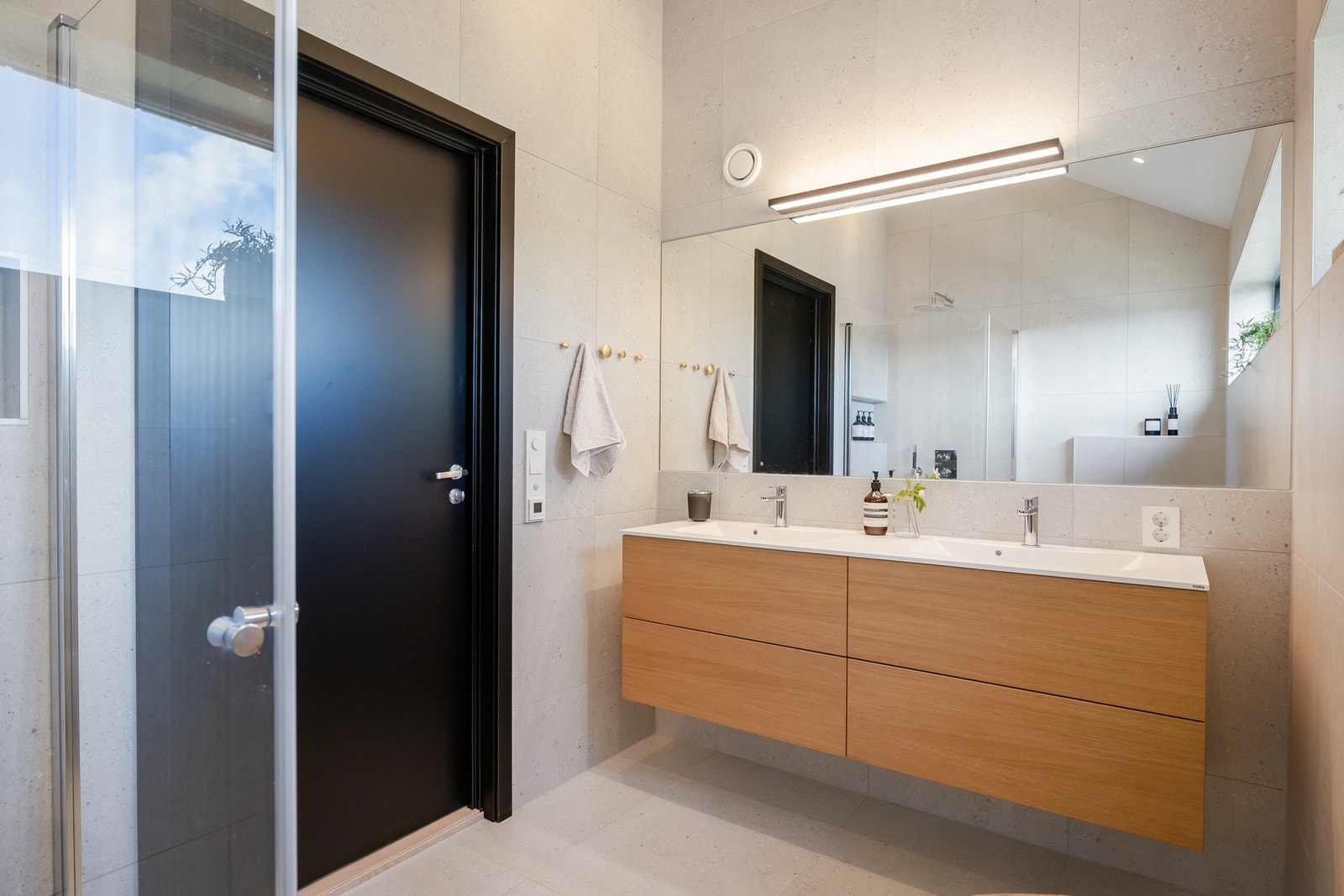 Lekkert baderom med flislagte overflater, innredning i type Eik Horisont fra Fossline med dobbel servant og innfelt speil fra vegg til vegg. Galleribilde