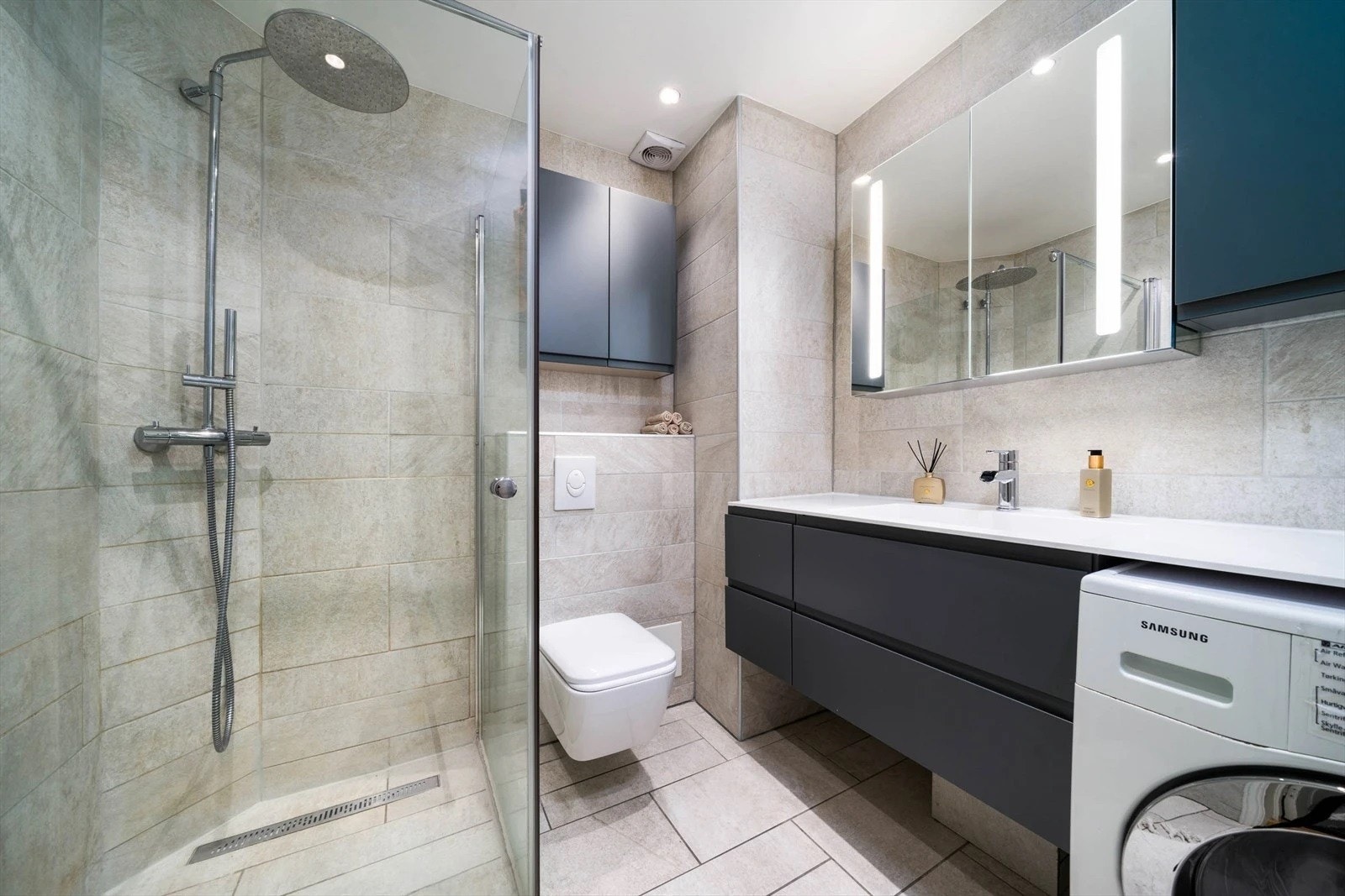 Pent flislagt baderom med romslig innredning med servant på underskap, speilskap med belysning, downlights, vegghengt wc, overskap og dusjhjørne med glassdører. Det er opplegg for vaskemaskin. Galleribilde