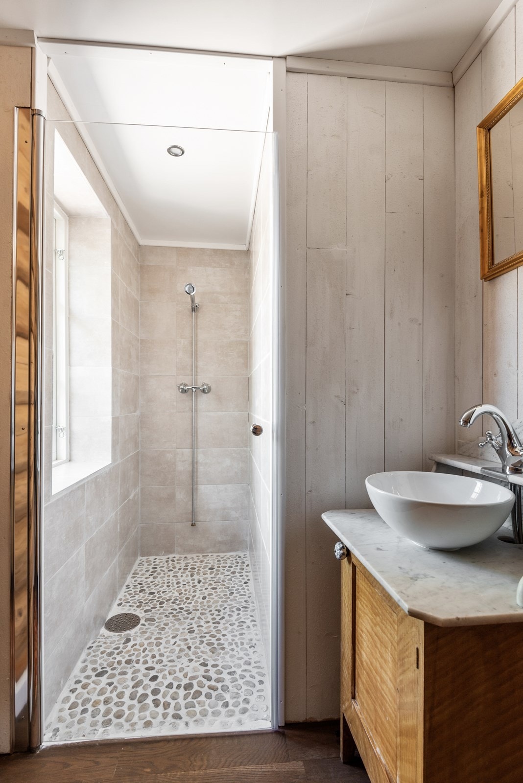 Pent sanitærrom med parkettgulv og vegger med tømmerpanel og malt panel. Dusjsone med rullestein på gulv og flislagte vegger. Innfelte downlights i himling. Innredninger med servant på underskap, forbrenningstoalett og vegghengt dusj i sone med glassdør. Galleribilde