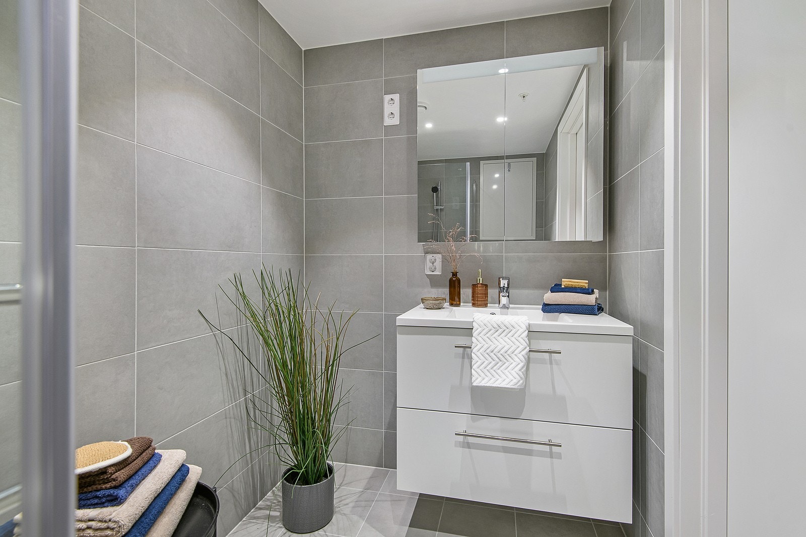 Lekkert flislagt baderom med varmekabler i gulv. Badet inneholder hvit baderomsinnredning, dusj med glassvegger, vegghengt toalett og opplegg for vaskemaskin. Galleribilde