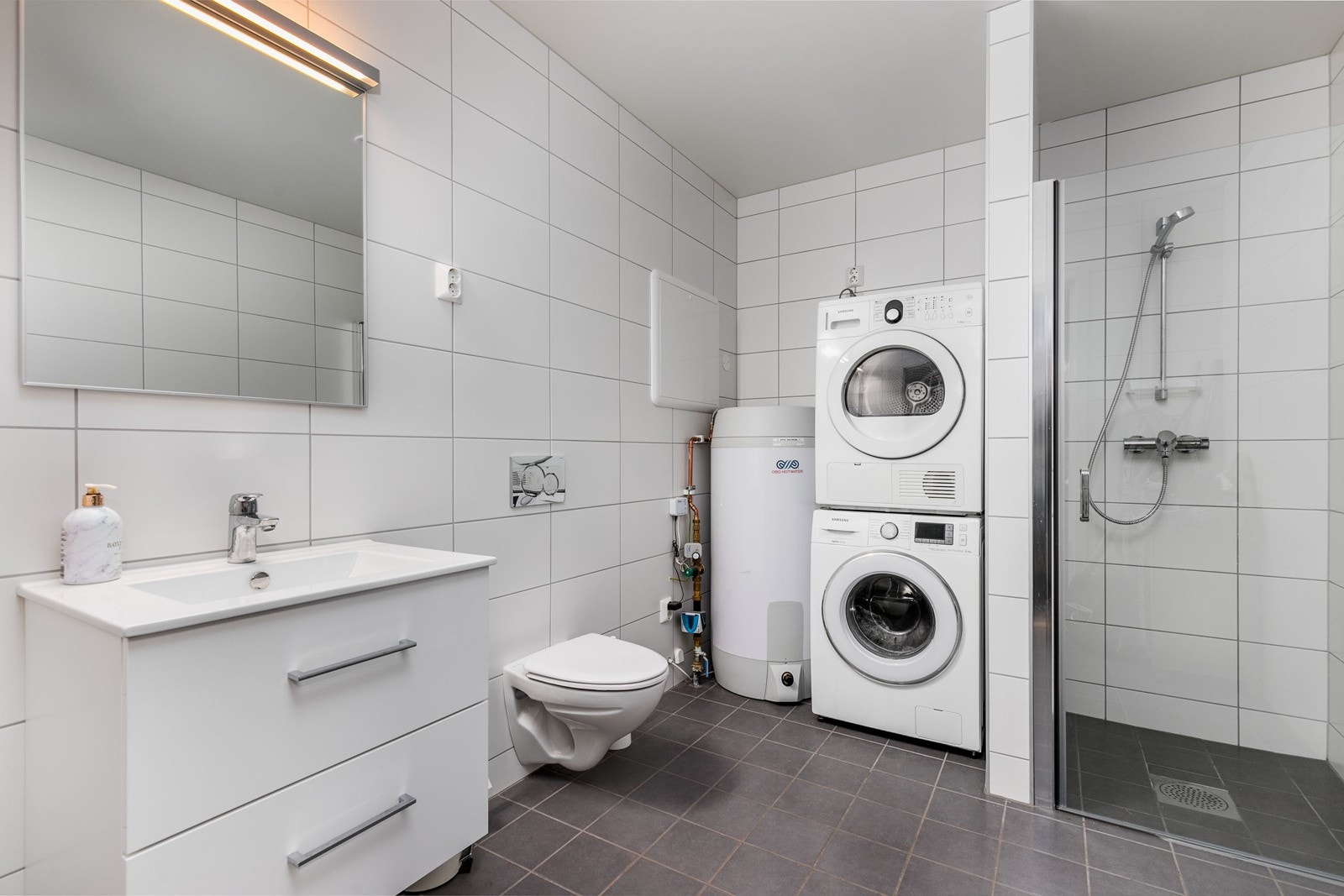 Flislagt bad i 1. etasje med varme i gulv. Innredningen består av servantskap og speil med belysning. Vegghengt klosett med innebygget sisterne. Dusjhjørne med dør. Opplegg til vaskemaskin og tørkerommel. Varmtvannsberederen er plassert her. Galleribilde