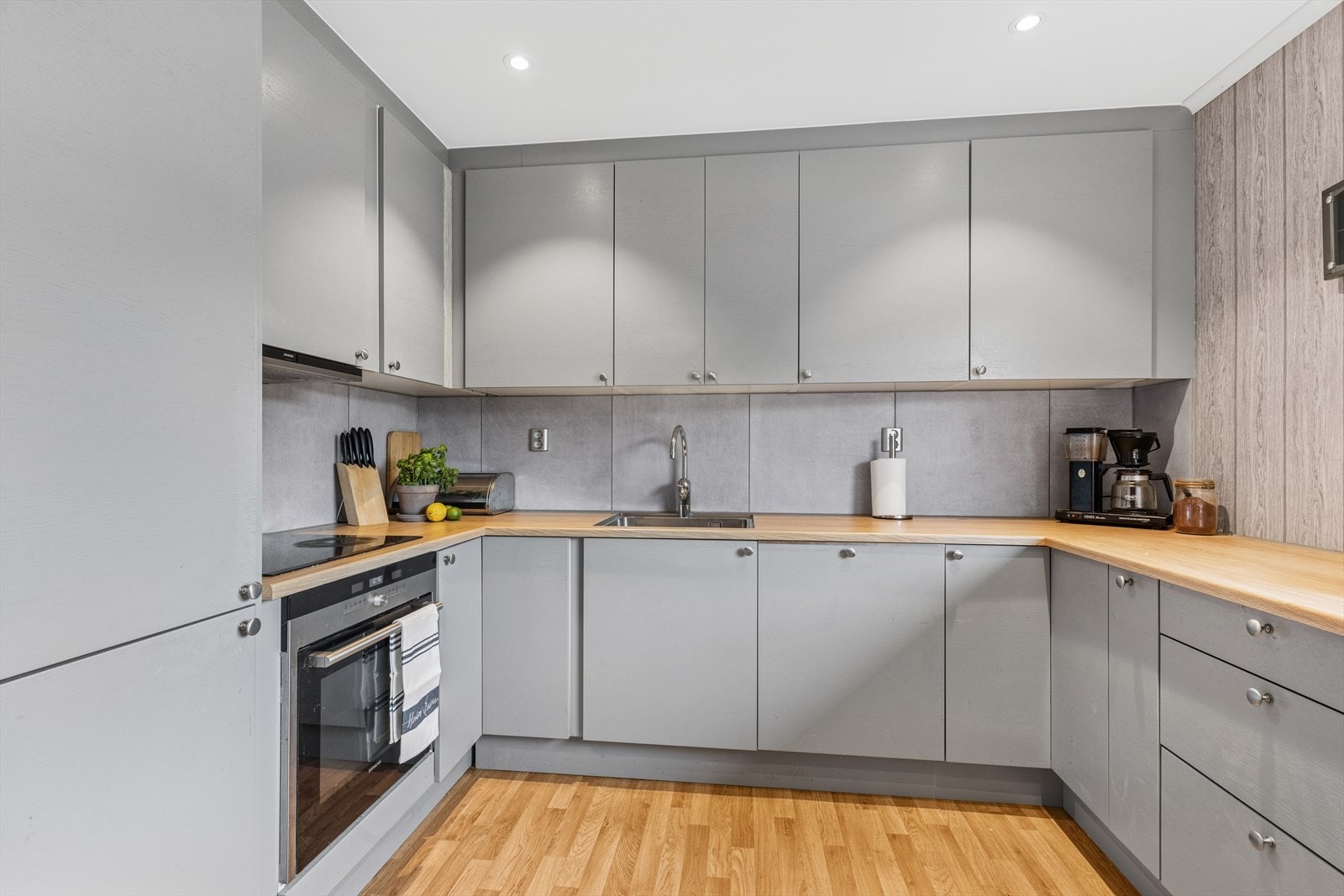 Kjøkkeninnredning fra Sigdal med grå fronter. Laminerte benkeplater med nedfelt oppvaskkum og fliser på vegg mellom overskap og kjøkkenbenk. Det er integrert kjøleskap med frysedel, oppvaskmaskin, komfyr og induksjonstopp. Galleribilde