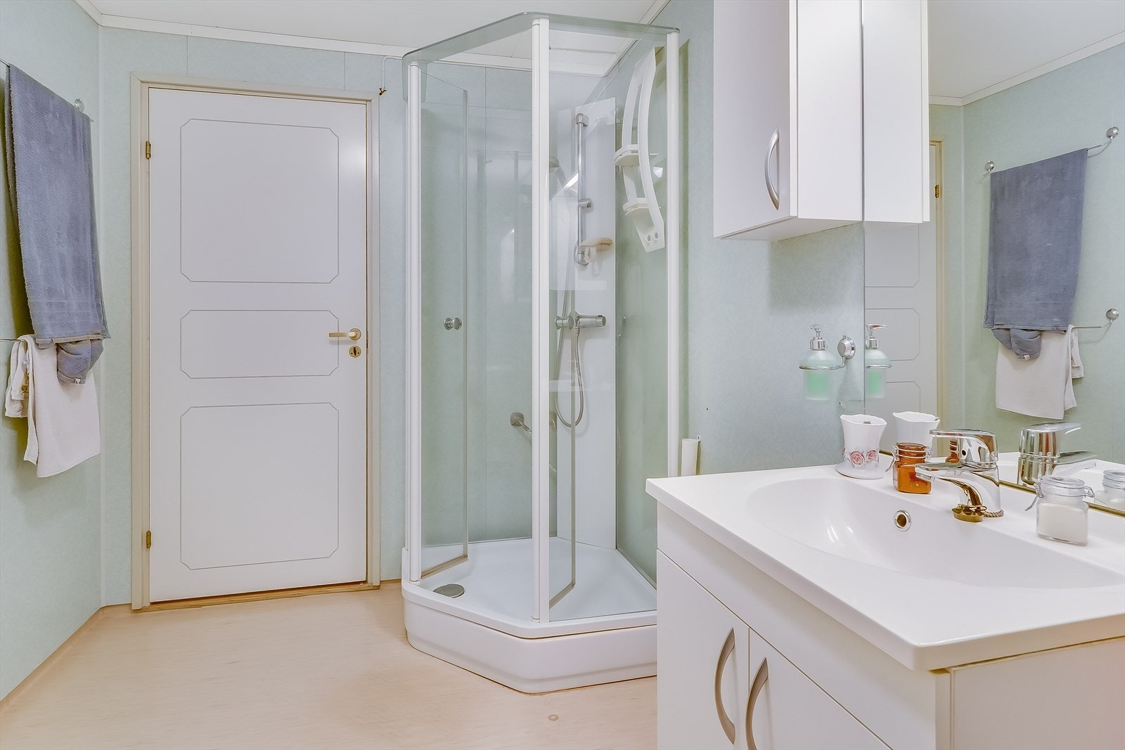 Badet er utstyrt med gulvmontert toalett, dusjkabinett og baderomsinnredning med underskap, heldekkende servant og veggskap. Opplegg for vaskemaskin. Galleribilde