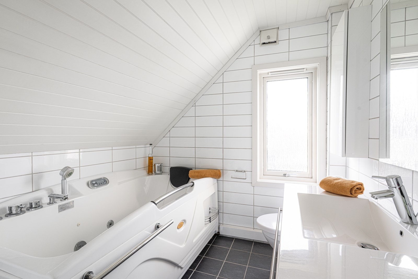 Flislagt bad med varmekabler i gulv. Badet er innredet med toalett, badekar og vegghengt servantskap med speil over. Galleribilde