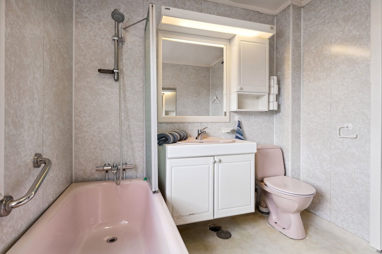 Lys baderomsinnredning med nedfelt rosa servant, speil med belysning og vegghengt skap over. Baderommet er også utstyrt med rosa badekar med dusjvegg, rosa gulvstående toalett og elektrisk stråleovn. Galleribilde