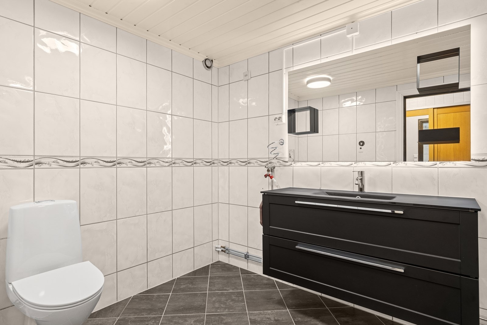 Badet er utstyrt med dusjkabinett, klosett, hel nedfelt servant i sort komposittmateriale, opplegg for vaskemaskin og baderomsinnredning med servantskap/skuffer, høyskap, speil og lys integrert bak speilet. Galleribilde