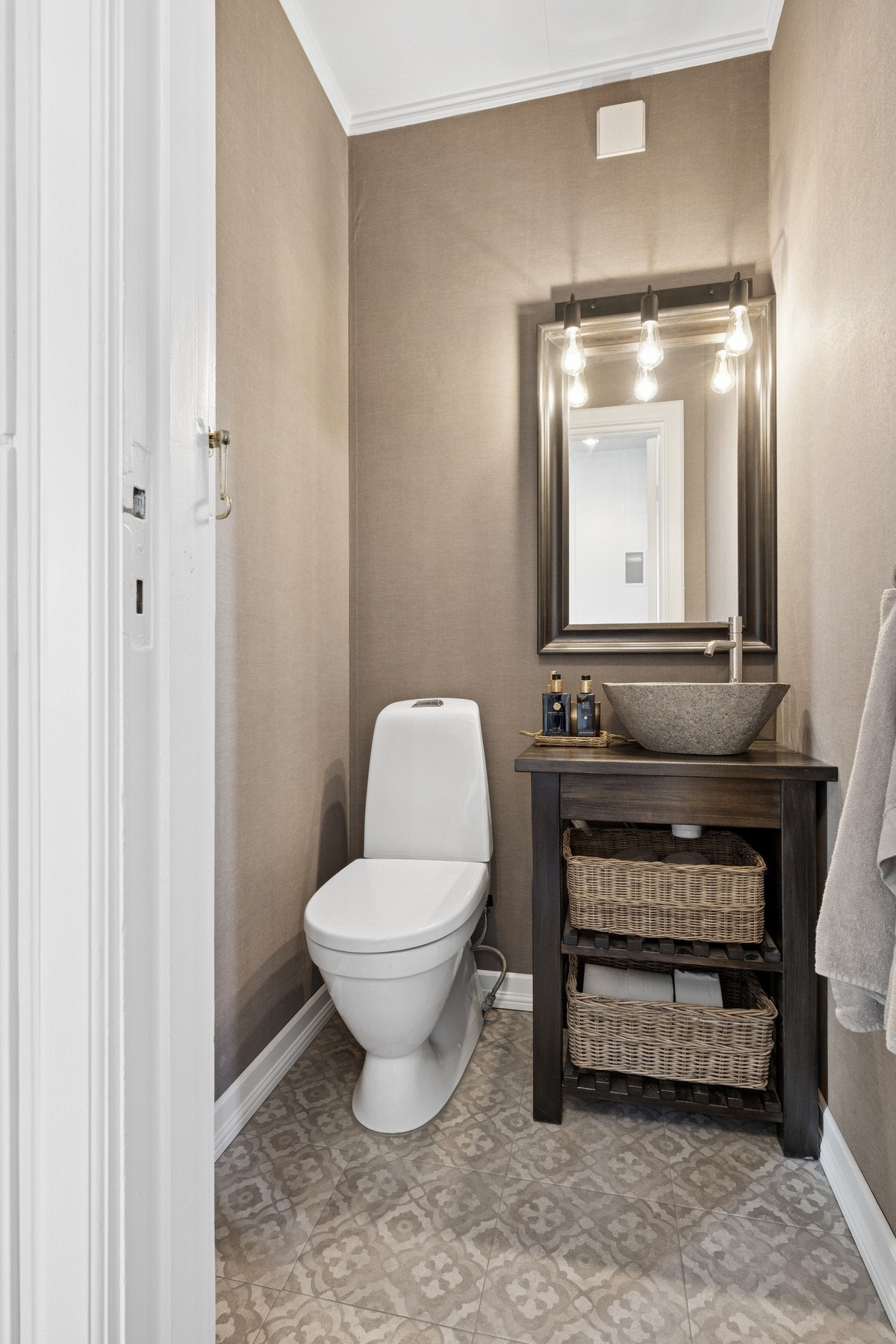 Pent toalettrom i lune farger med flislagt gulv, servant i naturstein og stilfull belysning - praktisk plassert rett ved inngangen. Galleribilde