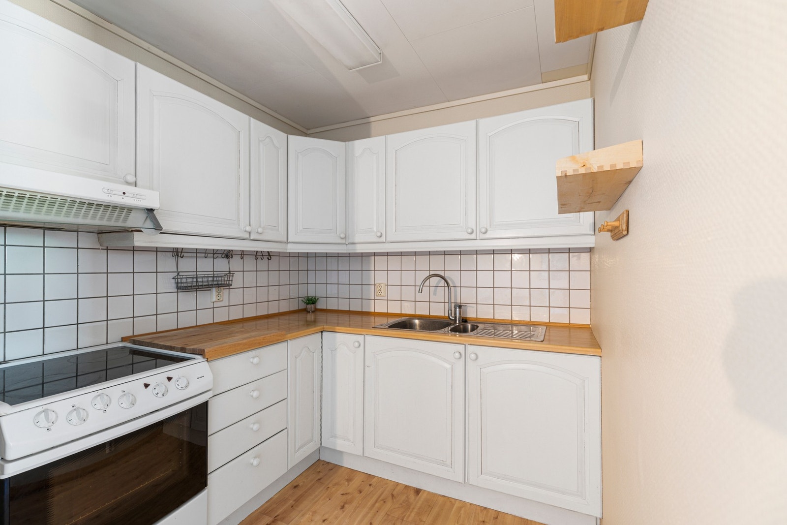 Kjøkkenet har en enkel innredning i hvite, profilerte fronter, laminat benkeplate med flis mot overskap, benkebeslag med oppvaskkum, ventilator og komfyr. Galleribilde