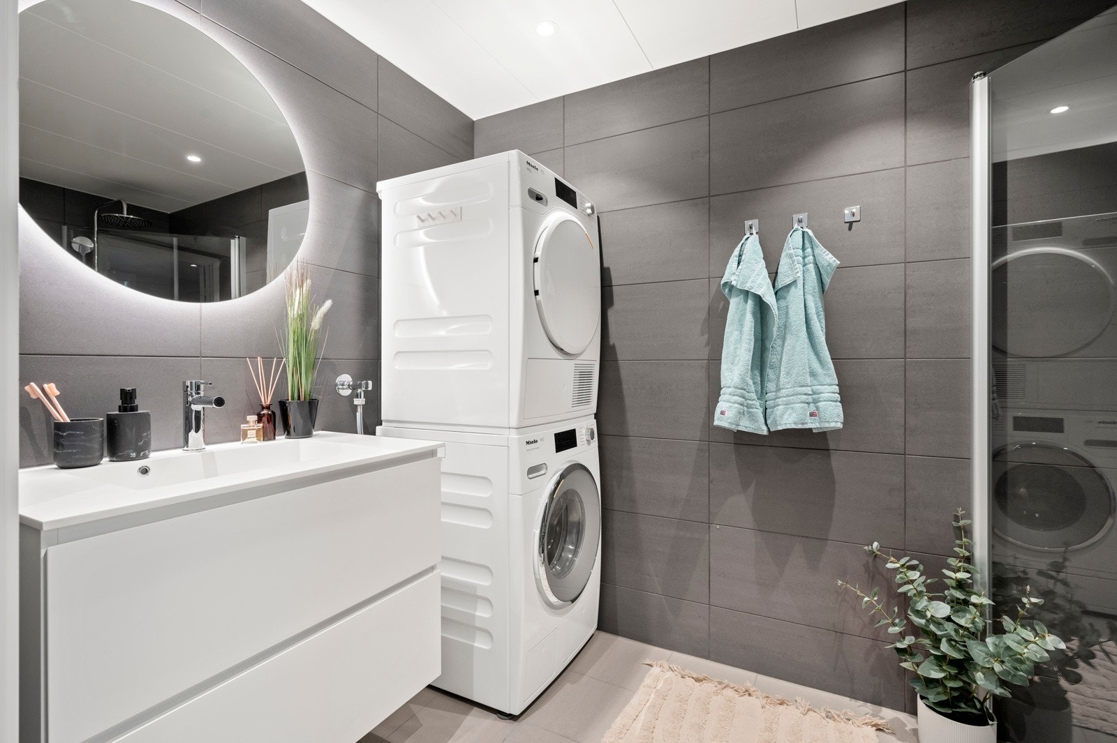 Dusjen er plassert på gulv og avskjermet med dusjdører i herdet glass. Videre er badet innredet med servant med skuffeinnredning, vegghengt toalett med innebygget sisterne, samt opplegg for både vaskemaskin og tørketrommel Galleribilde