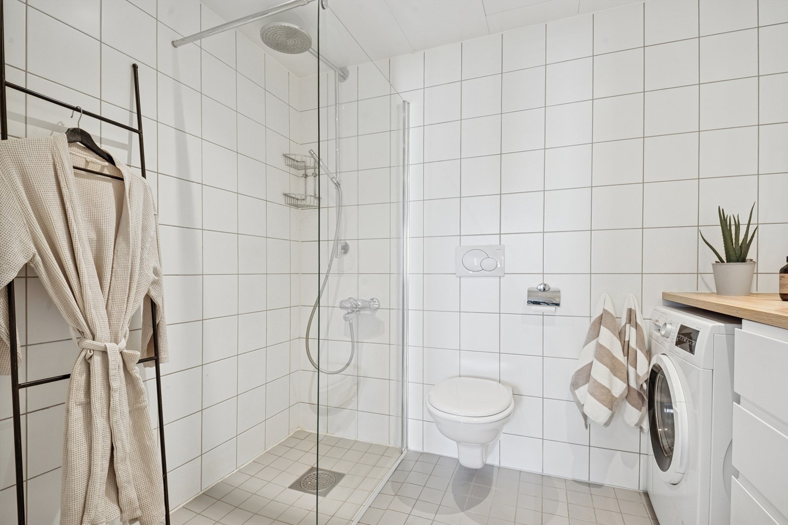 Rommet består av dusj på gulv med skillevegg i herdet glass, veggfestet klosett og servant med innfliset speil over. Rommet har opplegg for vaskemaskin. Benk- og overskap består av glatte fronter og bankeplaten er i tre. Galleribilde