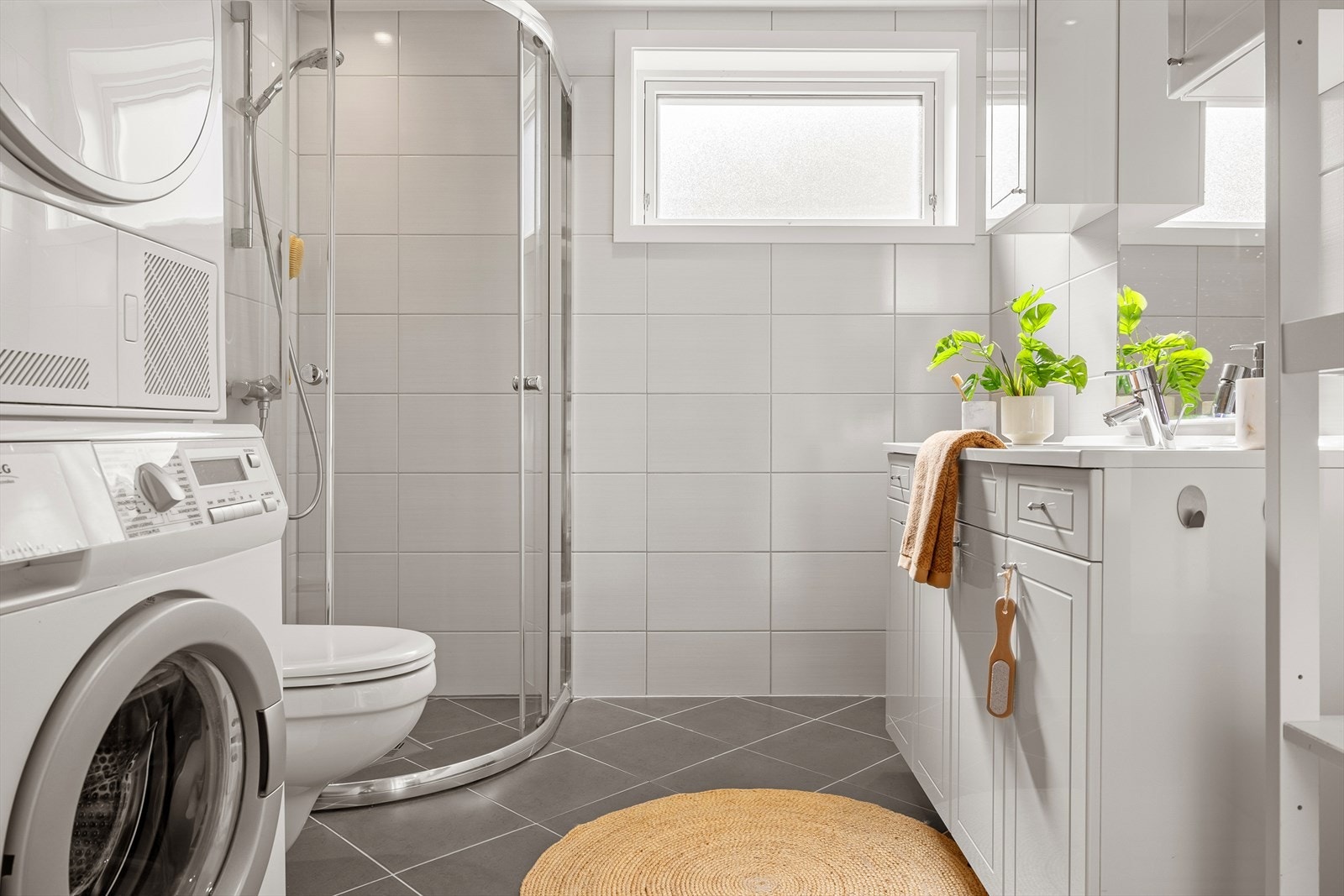 Badet har flislagte gulv og vegger, samt gulvvarme. Det er utstyrt med vegghengt toalett, helstøpt servant og underskap med profilerte fronter. Dusjen har dører i klart glass, og det er opplegg for vaskemaskin. Galleribilde