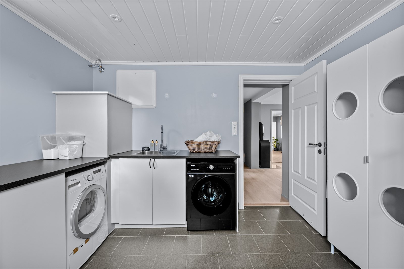 Stort og praktisk vaskerom med malte flater på veggene og fliser på gulv med gulvvarme. Vaskerommet har skapreoler med benkeplate og nedfelt utslagskum og opplegg for vaskemaskin og tørketrommel. Galleribilde