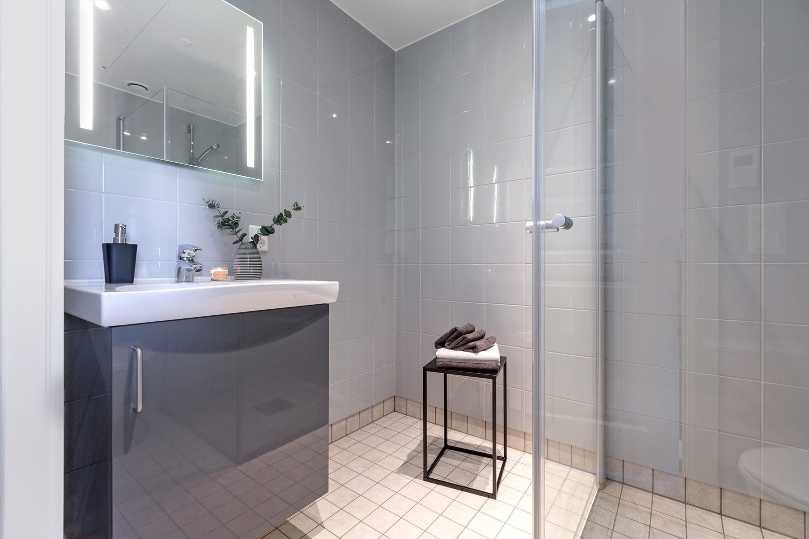 Badet har fliser på vegger og gulv med gulvvarme og er innredet med servant med skapseksjon, speil med tilhørende belysning, veggmontert toalett og dusjhjørne. Galleribilde