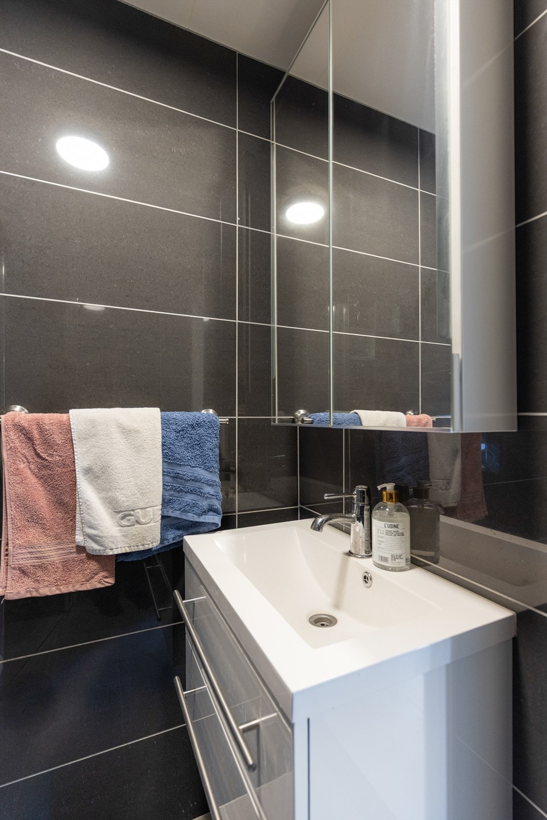 Badet inneholder veggmontert toalett, dusjnisje med glassdører og servantinnredning med underskuffer og vegghengt skap med speil. Galleribilde