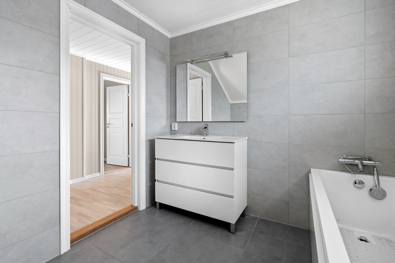 Rommet inneholder servant med underskap, speil med overbelysning, vegghengt toalett og badekar. Galleribilde