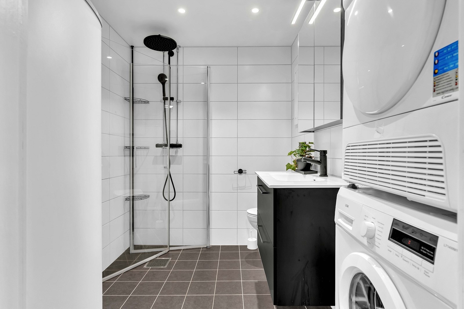 Delikat baderom med flislagte vegger og gulv. På badet er det dusjhjørne med innfellbare glassdører, vegghengt toalett, servantinnredning og opplegg for vaskesøyle. Galleribilde