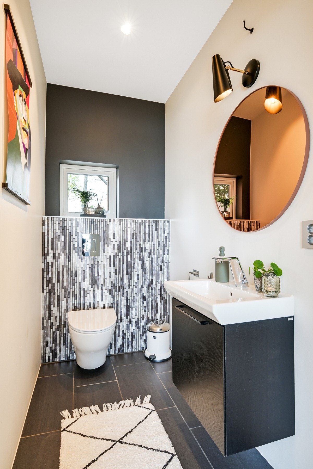 Wc-rommet har downlights, vegghengt toalett og flislagt gulv med varme. Et sort servantskap er plassert under et rundt speil med tilhørende lys Galleribilde