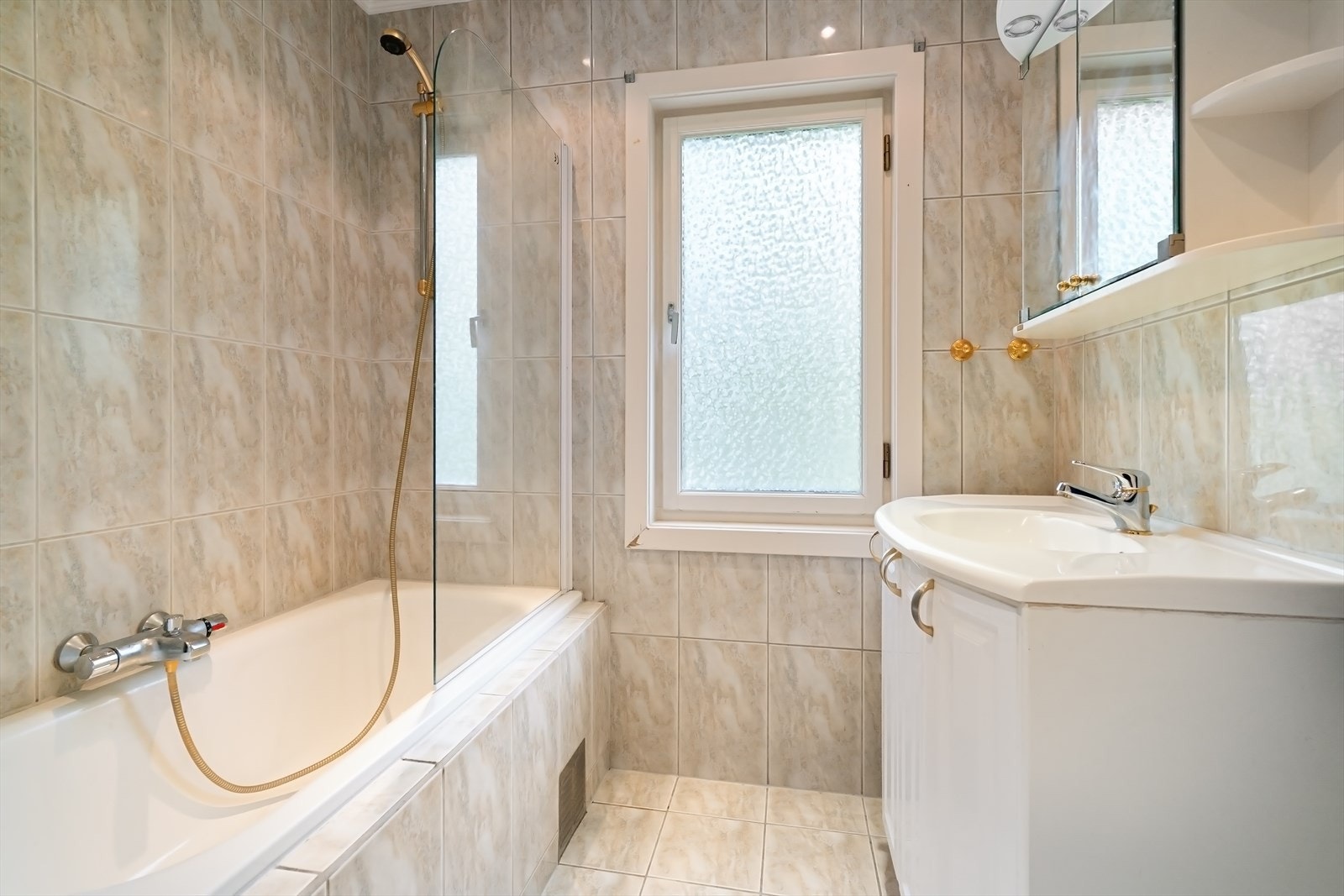 Pent baderom med flislagte gulv og vegger. Badet er utstyrt med toalett, servant med skap og innmurt badekar. Galleribilde