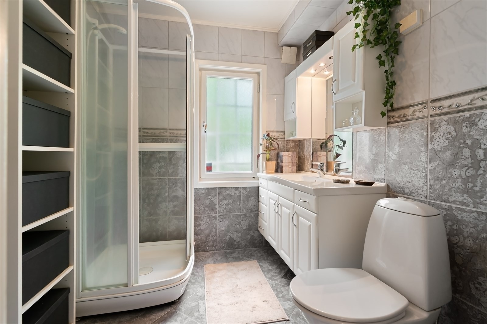 Baderom innredet med toalett, servant med skap og skuffer og dusjkabinett. Badet er flislagt med varmekabler i gulv og det er en fin vindusflate. Galleribilde