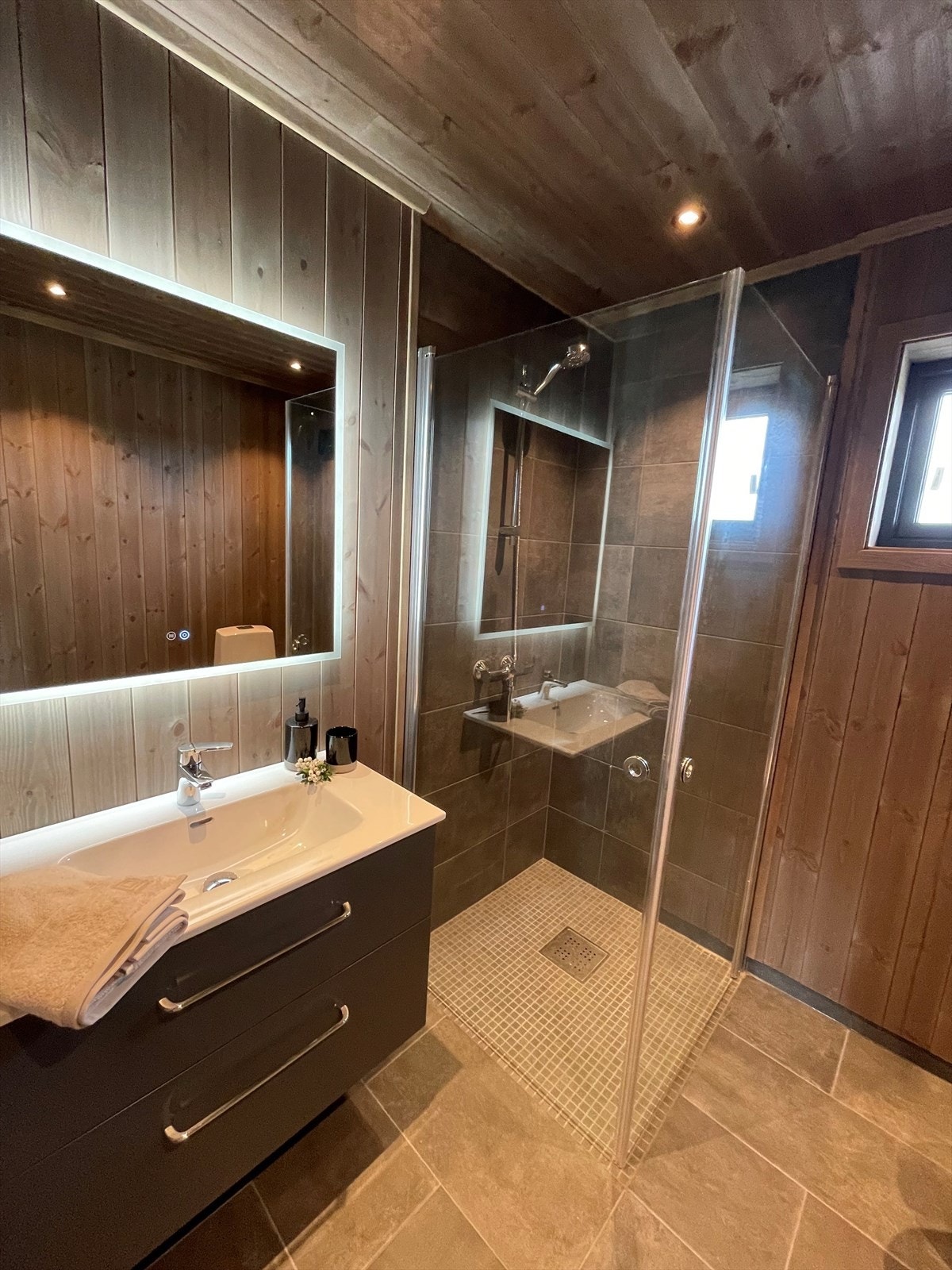 På badet er det stemningsfull downlightsbelysning og pent, flislagt gulv med varmekabler. Mørk baderomsinnredning med heldekkende servantplate i porselen og lyssatt speil over. Galleribilde