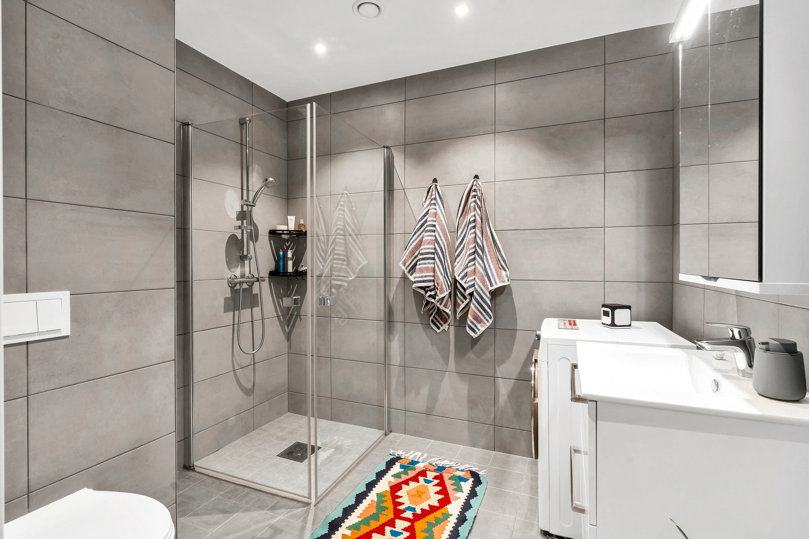 Bad med flislagt gulv og vegger, gulvvarme, speilskap, vegghengt servant med skuffer, opplegg for vaskemaskin og dusjhjørne med dører. Galleribilde